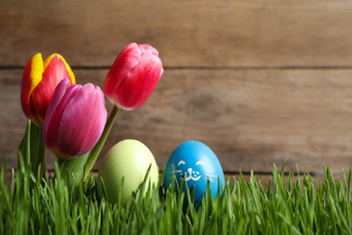 Photo of Colorful Easter eggs and tulip flowers in green grass against wooden background, closeup. Space for text