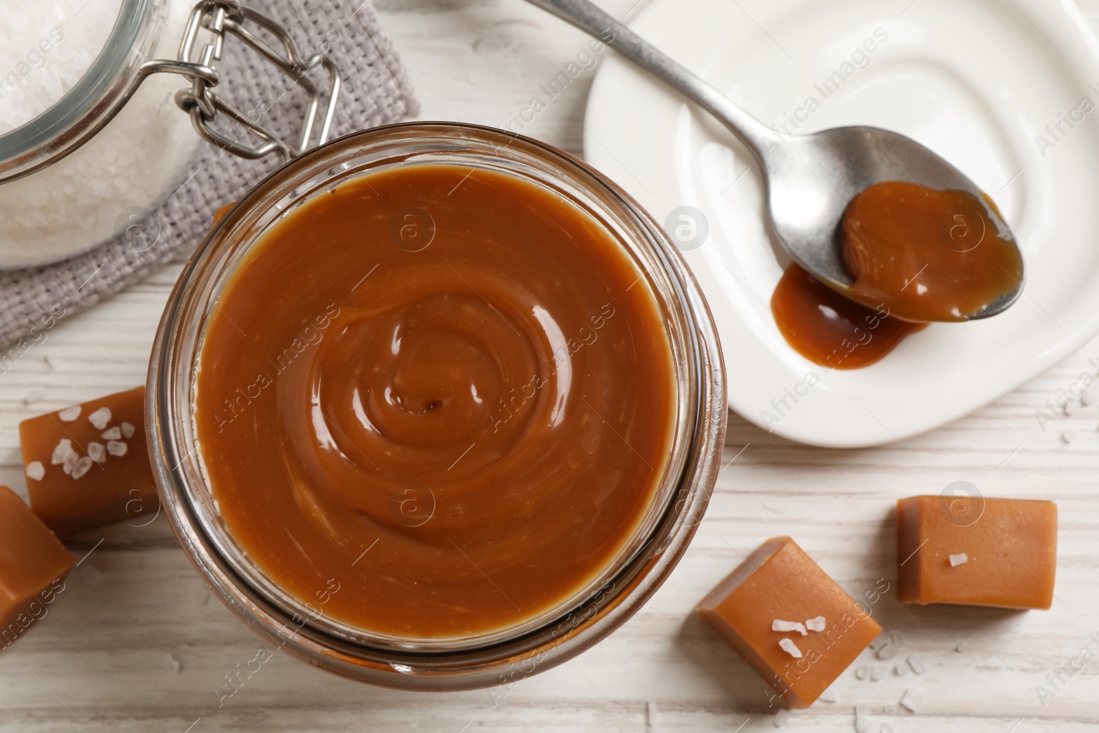 Photo of Yummy salted caramel in glass jar and candies on white wooden table, flat lay