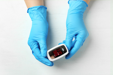 Photo of Doctor in latex gloves holding fingertip pulse oximeter on white wooden background, top view