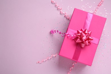 Pink gift box with confetti and streamers on lilac background, flat lay. Space for text