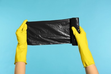 Photo of Janitor in rubber gloves holding roll of black garbage bags on light blue background, closeup