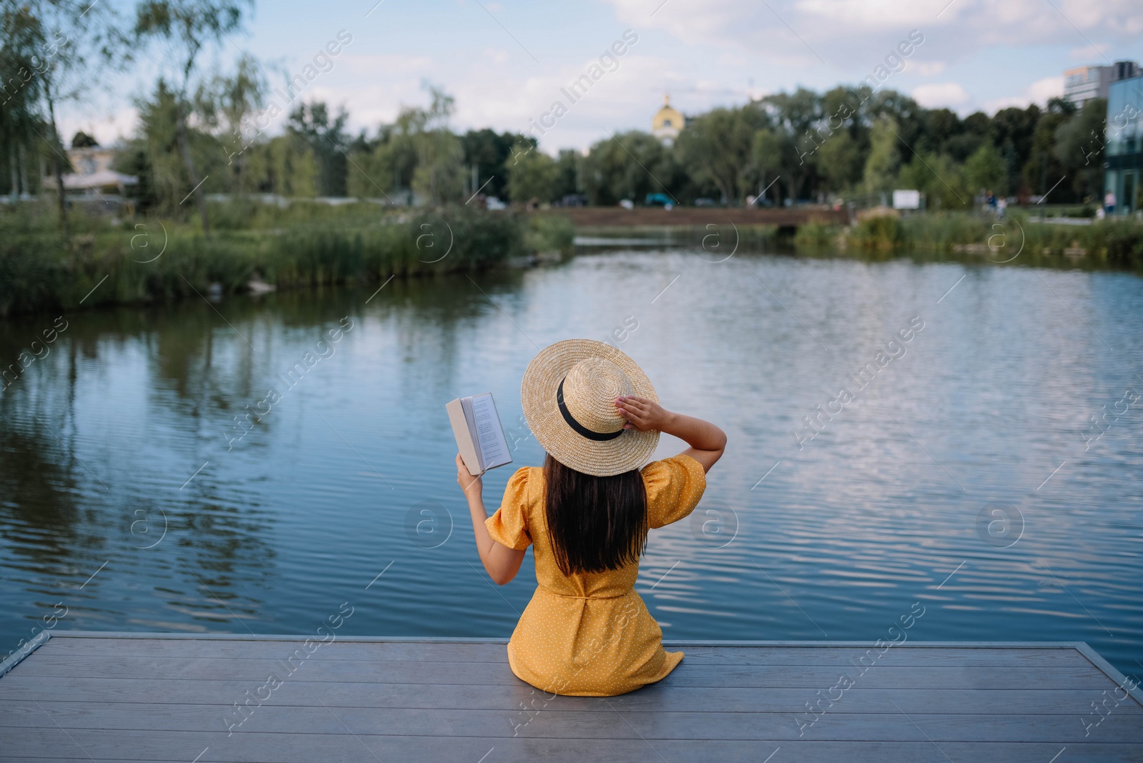 Photo of Woman reading book on pier near lake, back view