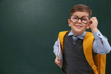 Photo of Cute little child wearing glasses near chalkboard, space for text. First time at school