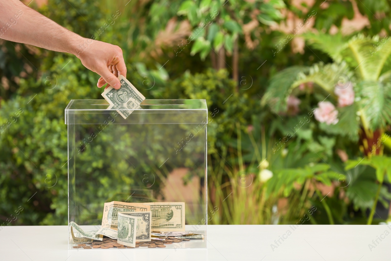 Photo of Man putting money into donation box on table against blurred background, closeup. Space for text