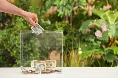 Photo of Man putting money into donation box on table against blurred background, closeup. Space for text