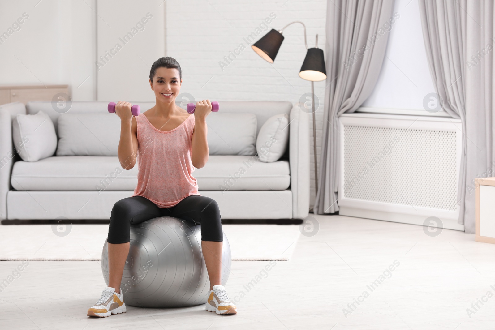 Photo of Young woman doing exercise with dumbbells on fitness ball at home. Space for text