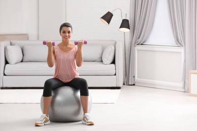 Photo of Young woman doing exercise with dumbbells on fitness ball at home. Space for text