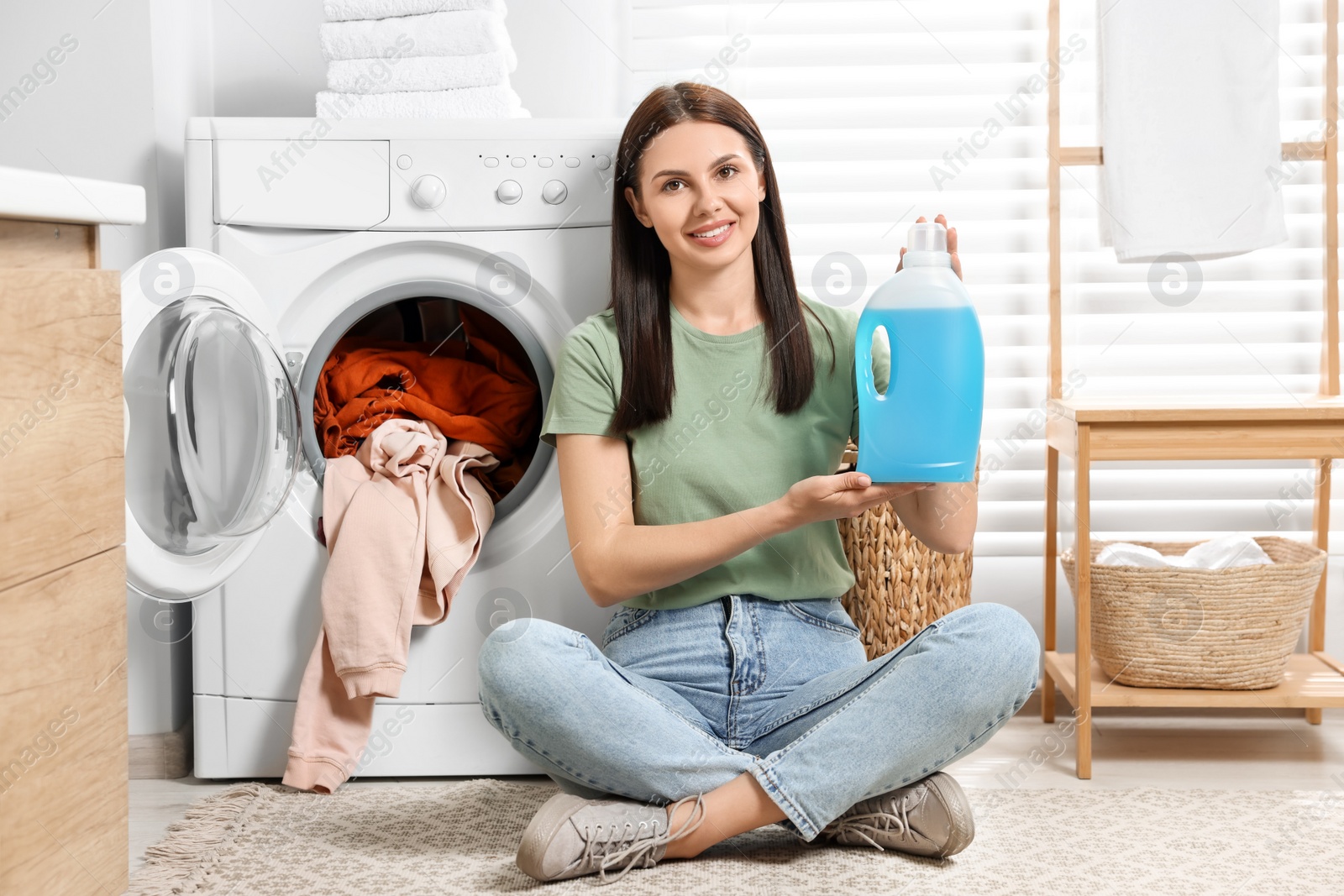 Photo of Woman sitting on floor near washing machine and holding fabric softener in bathroom