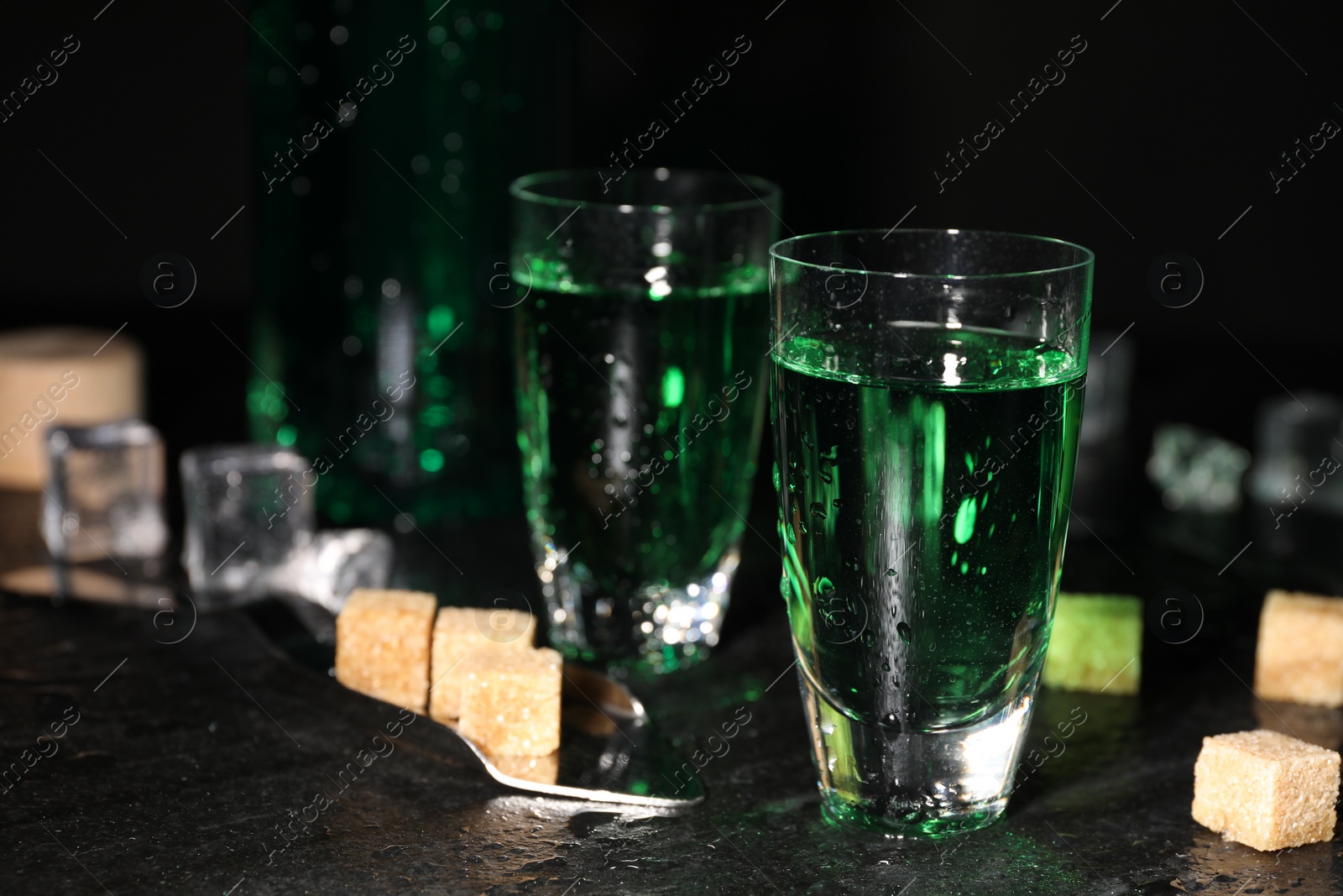 Photo of Absinthe in shot glasses, spoon, brown sugar and ice cubes on gray table, closeup. Alcoholic drink