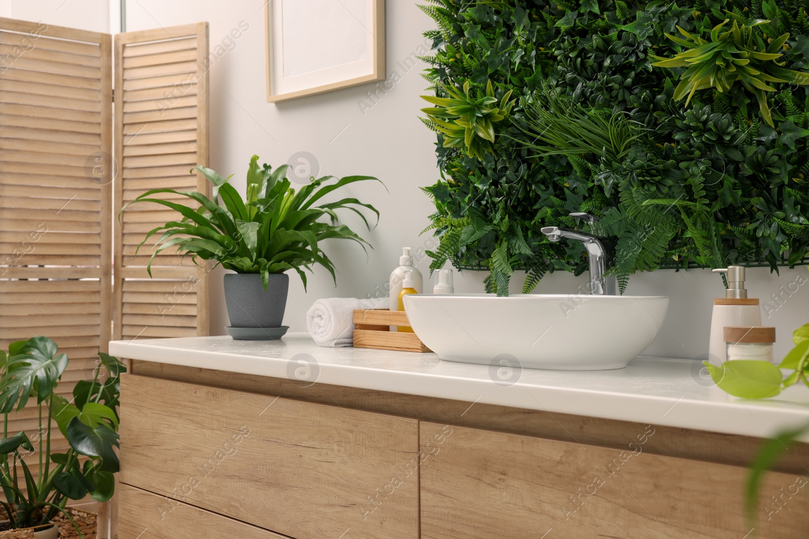 Photo of Green artificial plants, vessel sink and different personal care products in bathroom