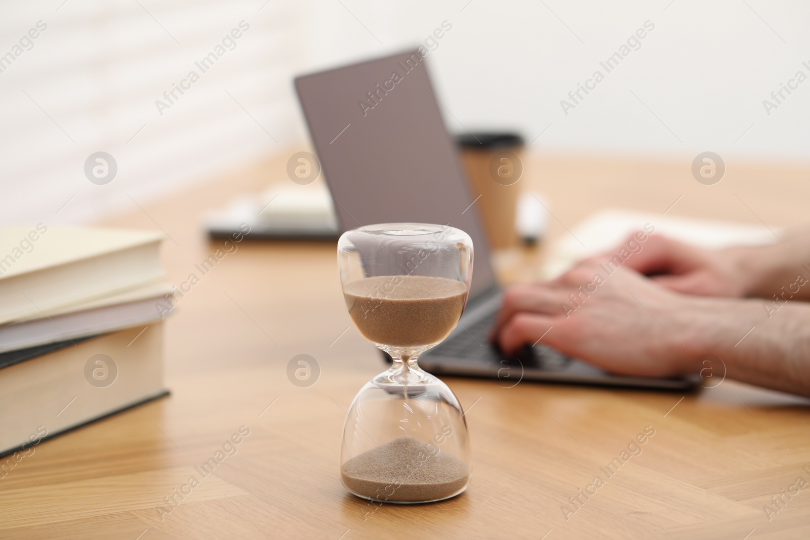 Photo of Hourglass with flowing sand on desk. Man using laptop indoors, selective focus