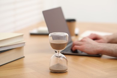 Hourglass with flowing sand on desk. Man using laptop indoors, selective focus