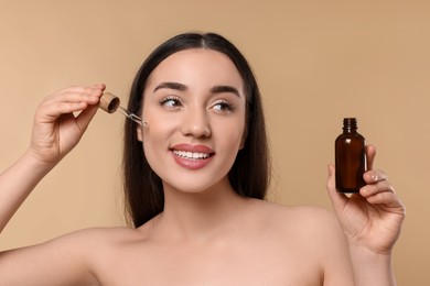 Photo of Happy young woman with bottle applying essential oil onto face on beige background