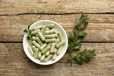 Photo of Vitamin capsules in bowl and leaves on wooden table, top view