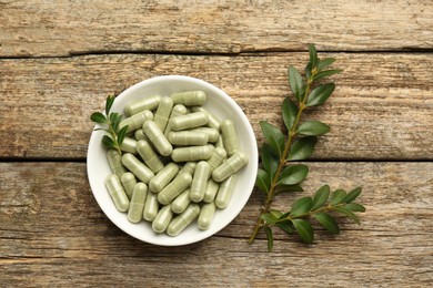 Vitamin capsules in bowl and leaves on wooden table, top view