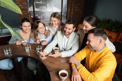 Photo of Handsome man showing something funny in smartphone to his friends in cafe
