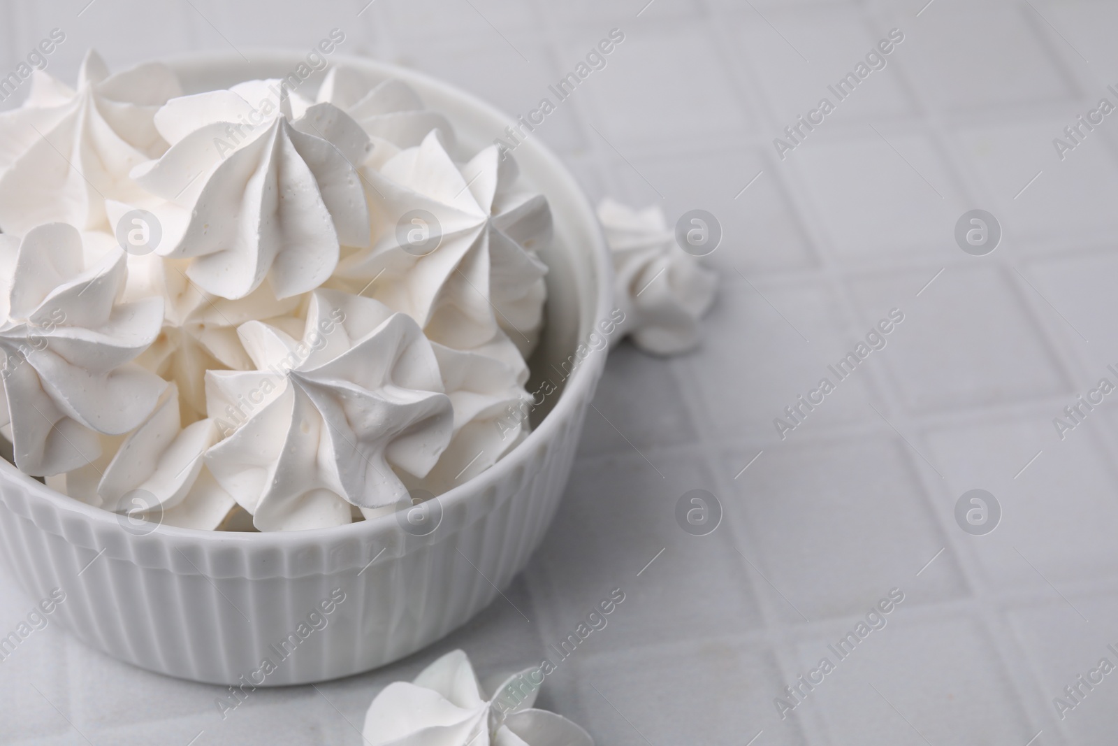 Photo of Tasty meringue cookies in bowl on white tiled table. Space for text