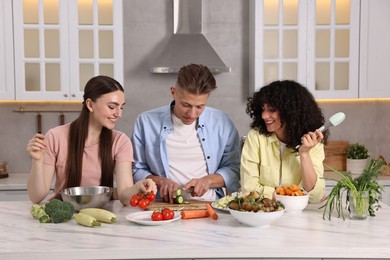 Photo of Friends cooking healthy vegetarian meal at white marble table in kitchen