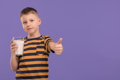 Photo of Cute boy with glass of fresh milk showing thumb up on violet background, space for text