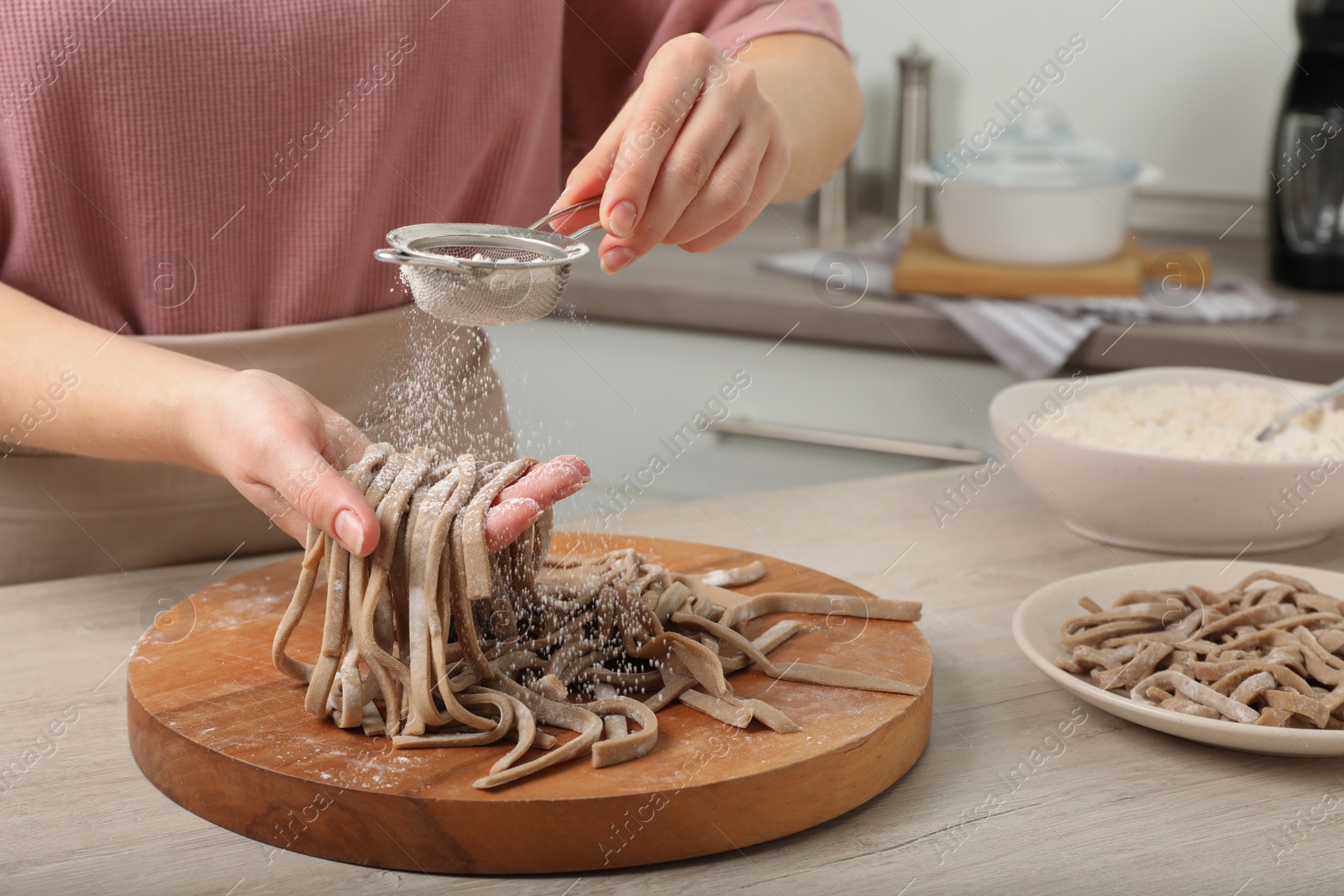 Photo of Woman sprinkling raw soba (buckwheat noodles) with flour at wooden table in kitchen, closeup