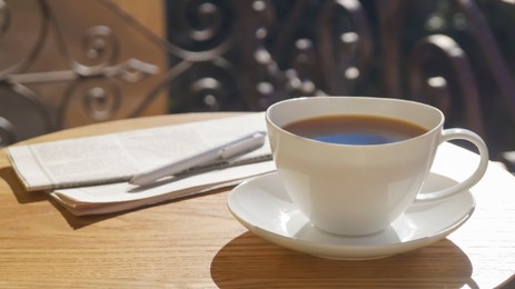 Cup of aromatic coffee, pen and newspaper on wooden table outdoors, space for text