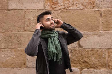 Photo of Smiling man in warm scarf listening to music near brick wall