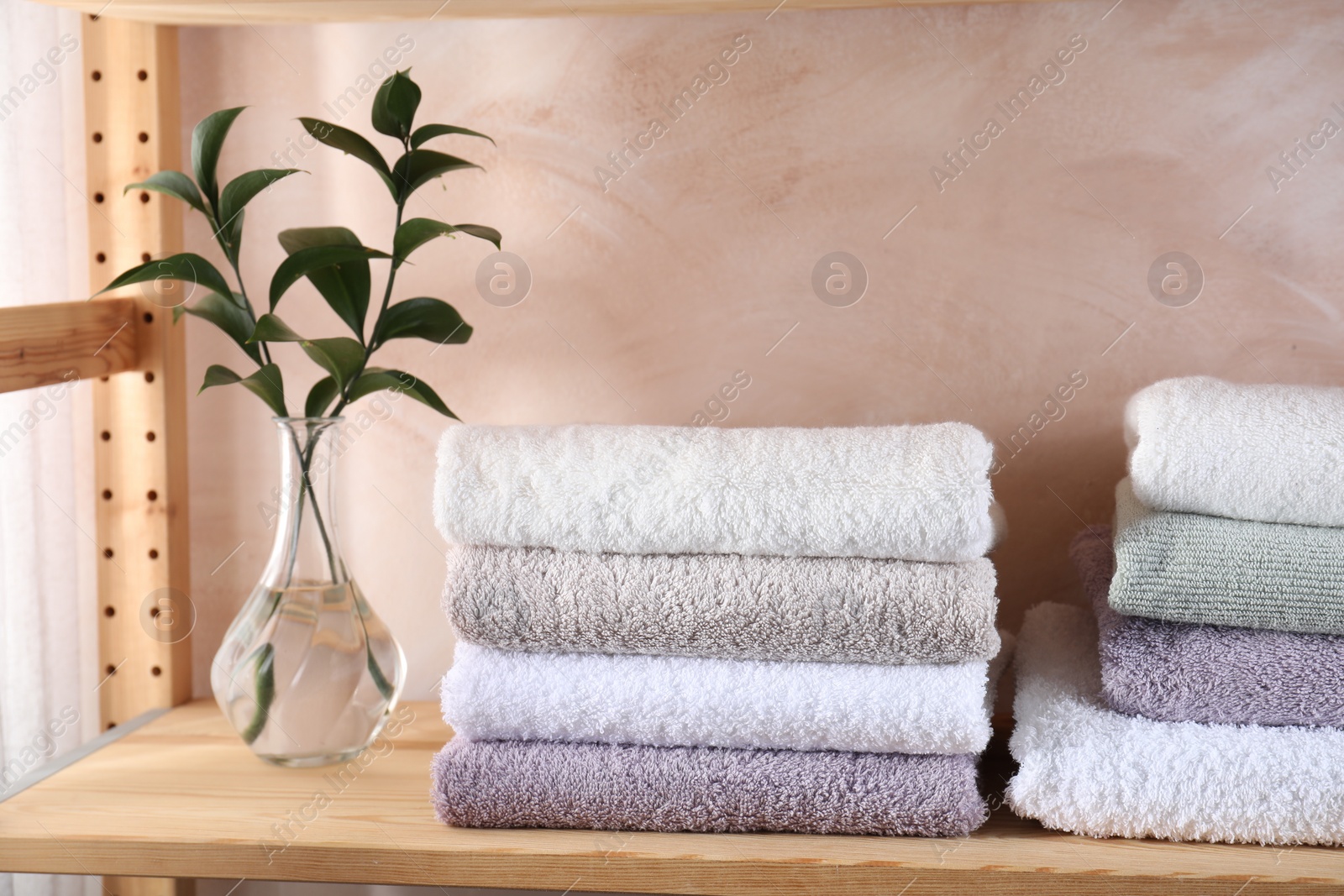 Photo of Stacked soft towels and green leaves on wooden shelf indoors