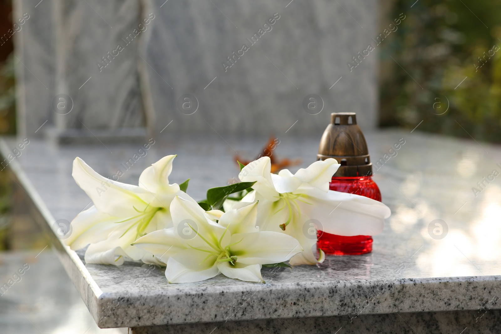 Photo of White lilies and grave light on granite tombstone outdoors. Funeral ceremony