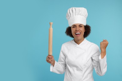 Photo of Emotional female chef in uniform holding rolling pin on light blue background. Space for text