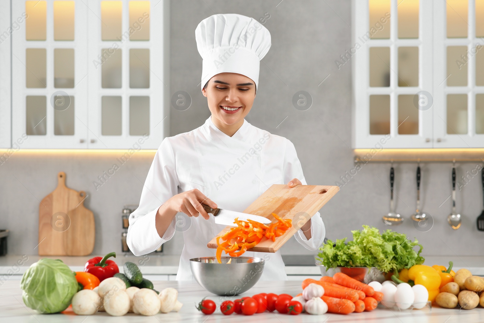 Photo of Professional chef putting cut bell pepper into metal bowl at white marble table in kitchen