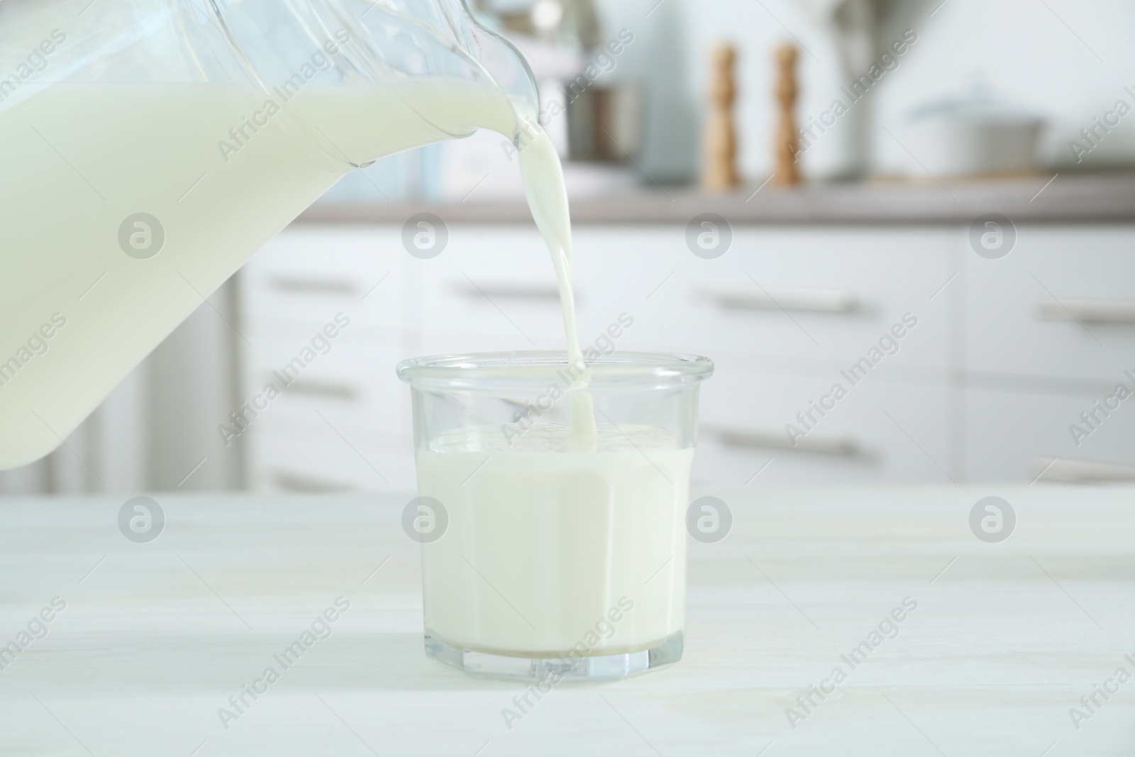 Photo of Pouring milk from jug into glass on white wooden table in kitchen