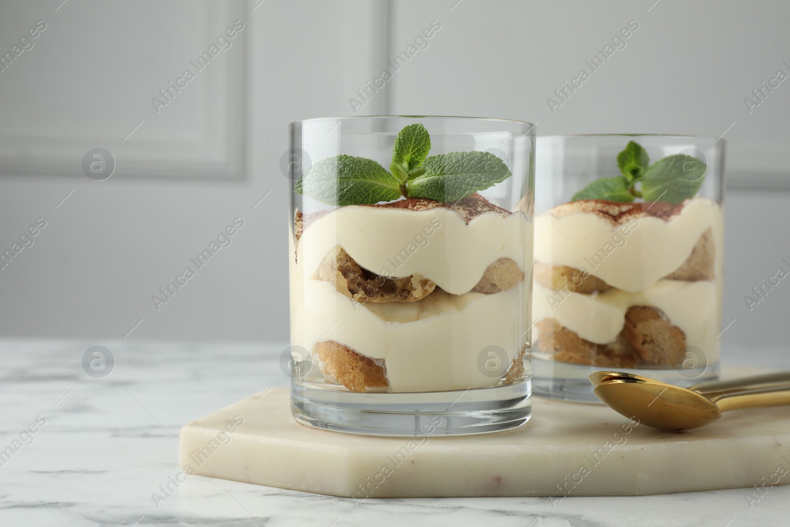 Photo of Delicious tiramisu in glasses, mint leaves and spoons on white marble table, space for text