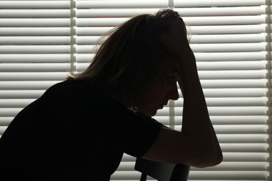 Photo of Silhouette of sad young woman near closed blinds indoors