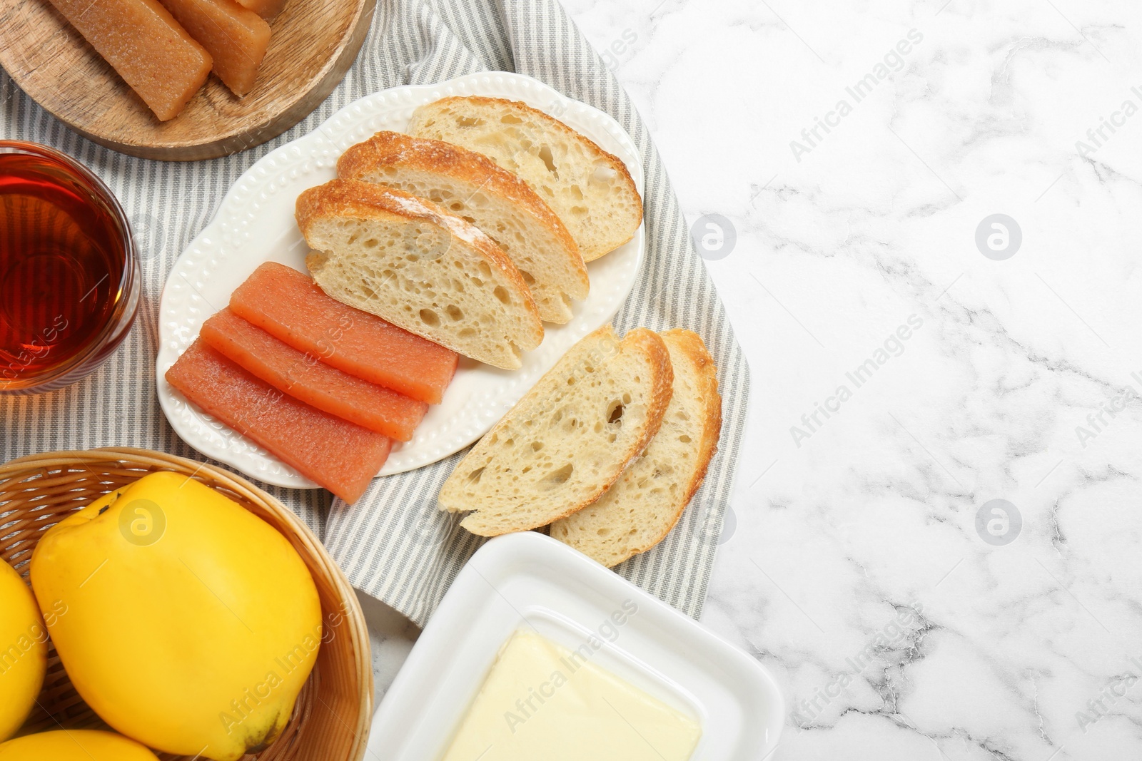 Photo of Delicious quince paste, bread, butter, cup of tea and fresh fruits on white marble table, flat lay. Space for text