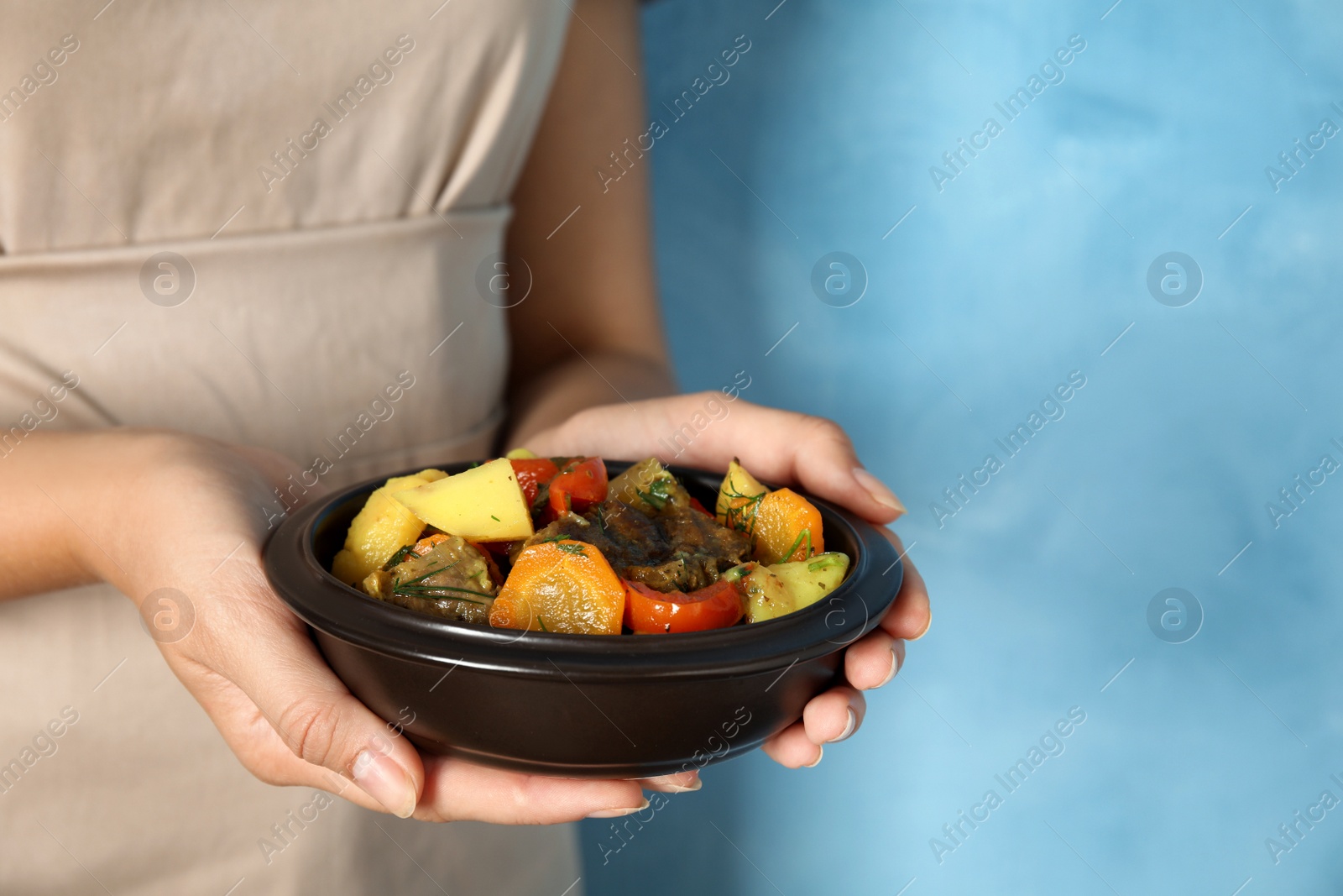 Photo of Woman holding earthenware with delicious roasted potatoes and meat on light blue background, closeup