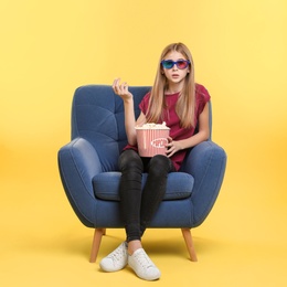 Emotional teenage girl with 3D glasses and popcorn sitting in armchair during cinema show on color background