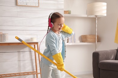 Photo of Cute little girl in headphones with mop singing while cleaning at home
