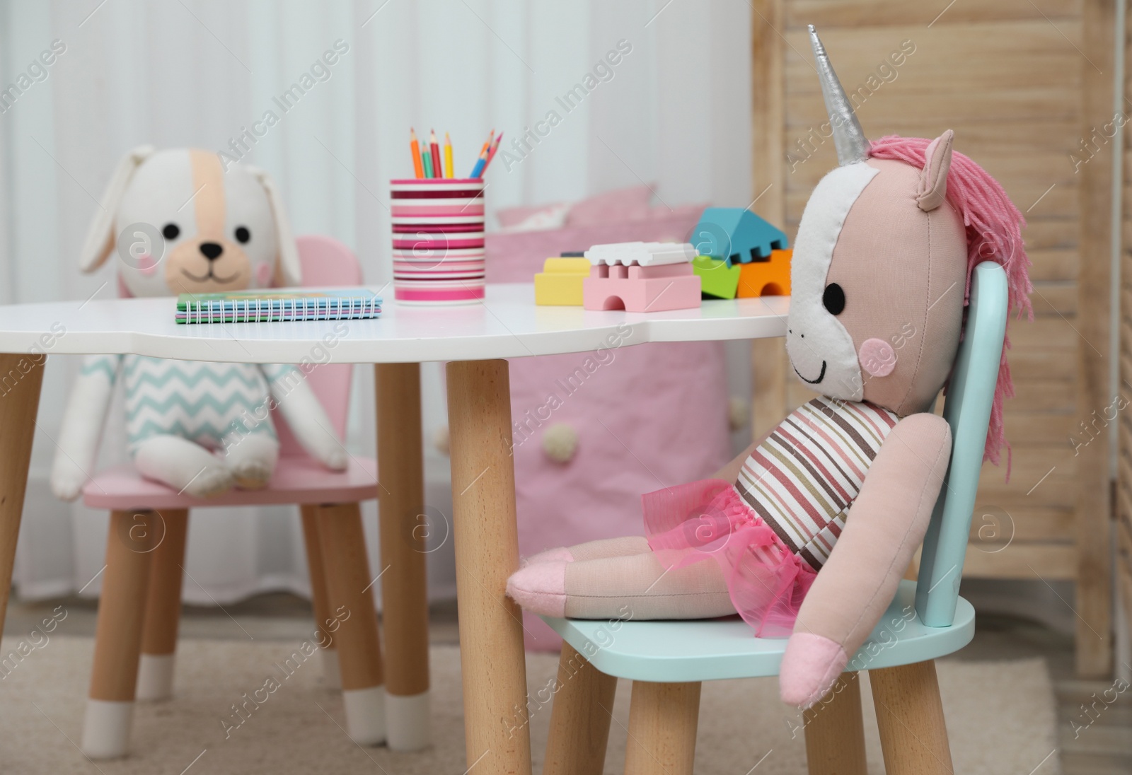 Photo of Cute toy unicorn and bunny on small chairs at table in playroom. Interior design