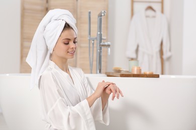 Photo of Beautiful woman in white robe applying cream near tub in bathroom