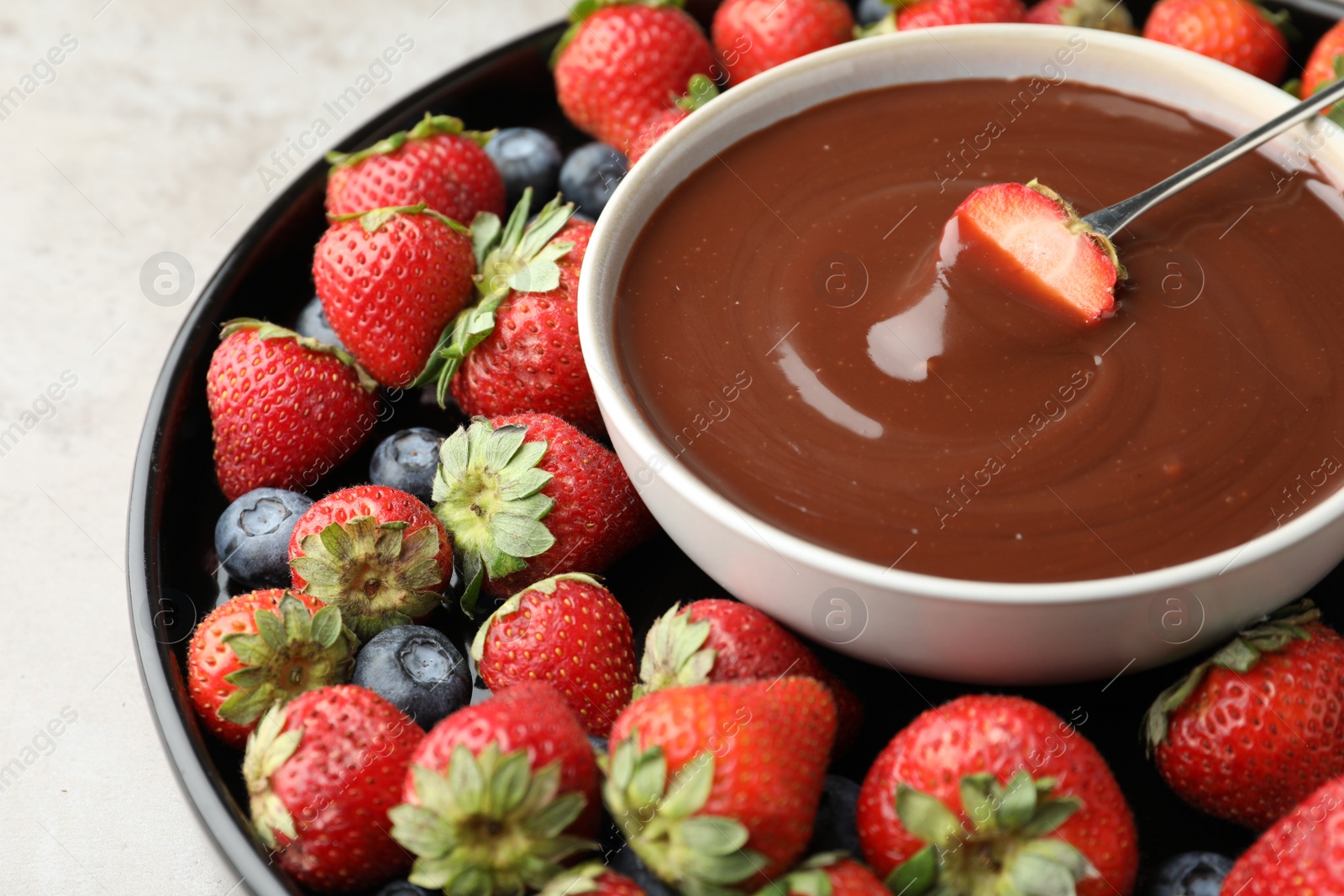 Photo of Fondue fork with strawberry in bowl of melted chocolate surrounded by different berries on light table, closeup