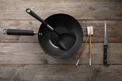 Photo of Black metal wok, chopsticks, knife and spatula on wooden table, flat lay