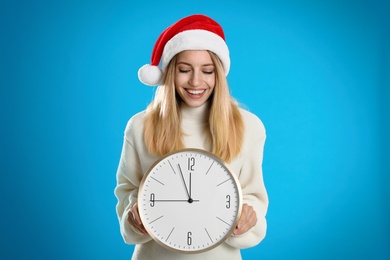 Woman in Santa hat with clock on light blue background. New Year countdown