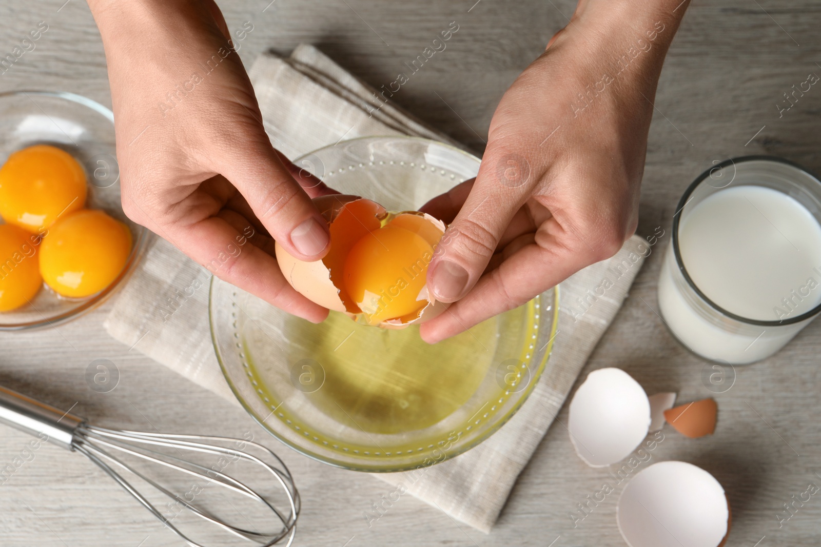 Photo of Woman separating egg yolk from white over glass bowl at light wooden table, top view
