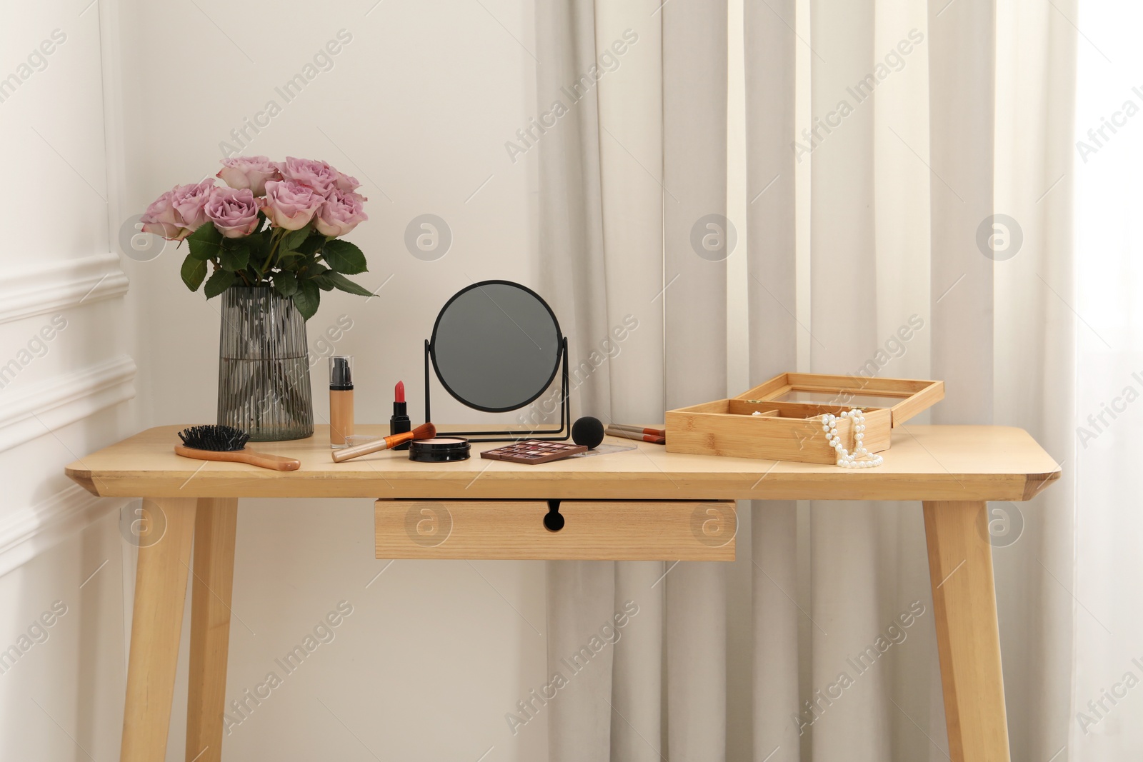 Photo of Mirror, cosmetic products, box of jewelry and vase with pink roses on wooden dressing table in makeup room