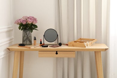 Mirror, cosmetic products, box of jewelry and vase with pink roses on wooden dressing table in makeup room
