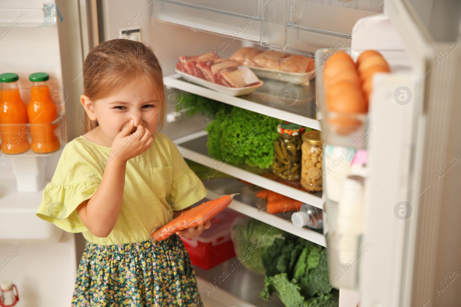 Photo of Cute little girl with bad smelling sausages near refrigerator at home