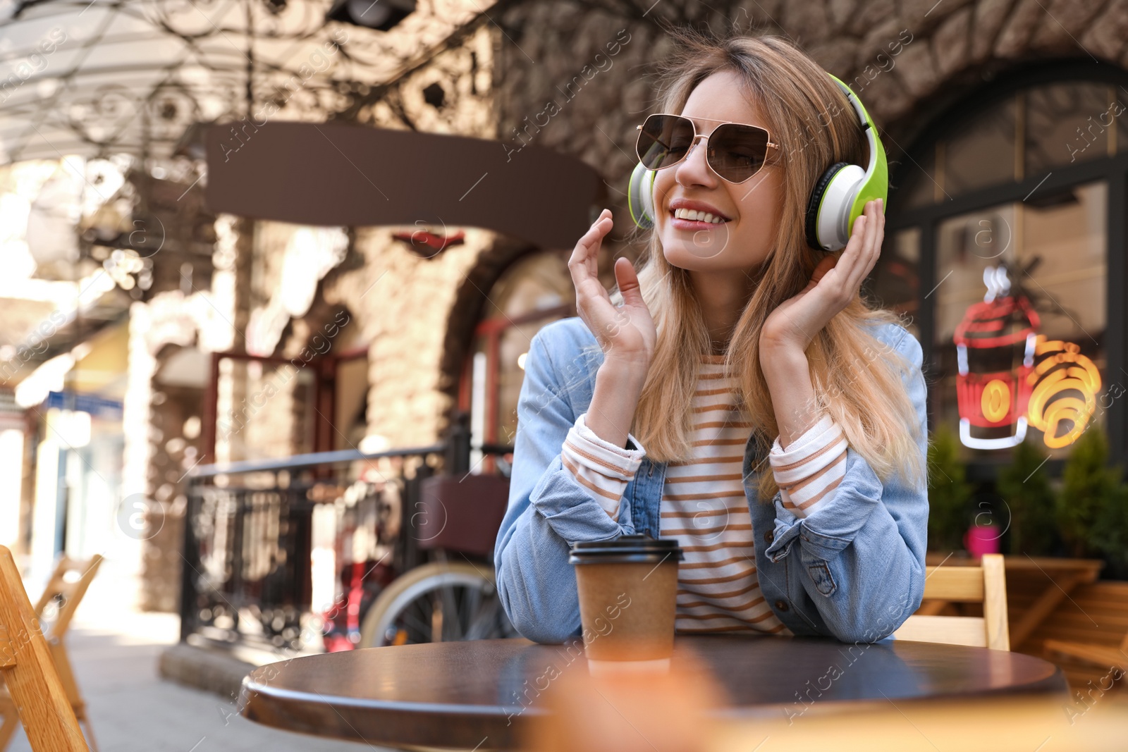 Photo of Happy young woman with coffee and headphones listening to music in outdoor cafe