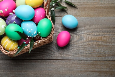 Colorful Easter eggs in wicker basket on wooden table, above view. Space for text