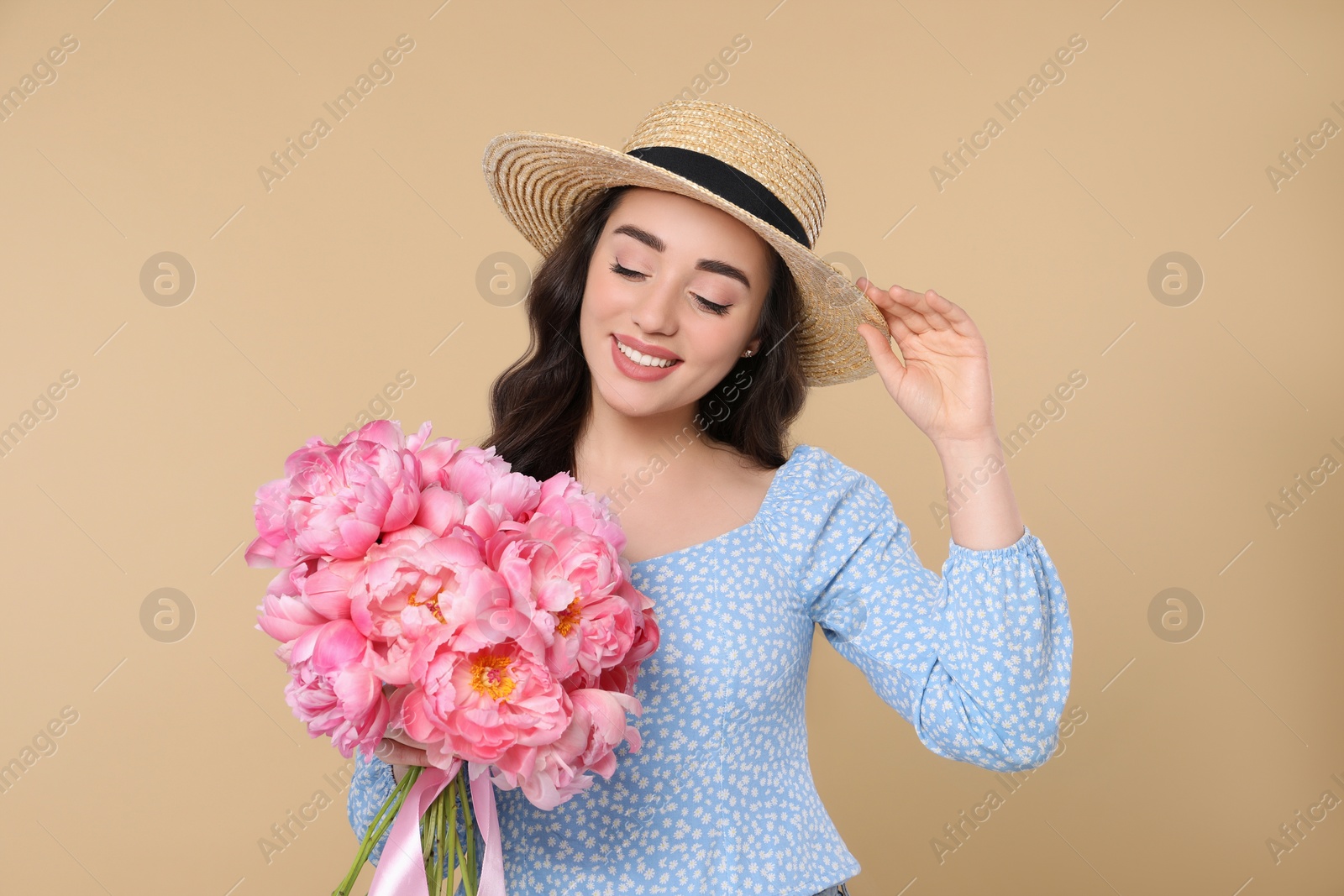 Photo of Beautiful young woman in straw hat with bouquet of pink peonies against beige background