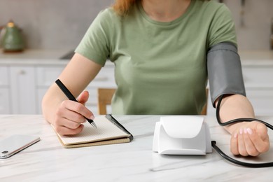 Photo of Woman measuring blood pressure and writing it down into notebook in kitchen, closeup